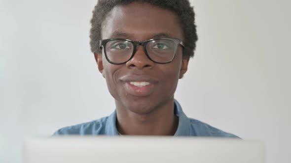 Close Up of Young African Man Smiling at Camera While Using Laptop in Office