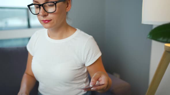 Woman with Glasses Sits on a Sofa in a Cozy Room and Makes an Online Shopping Using a Credit Card