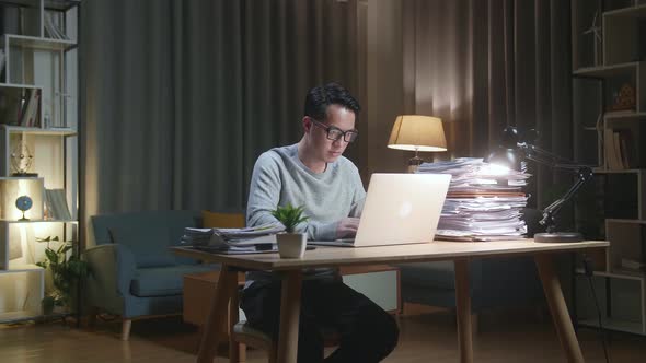 Asian Man Typing Laptop Computer While Working With Documents At Home