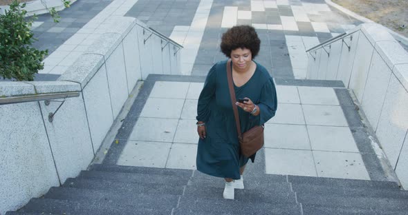 Plus size biracial woman walking on stairs with smartphone in city