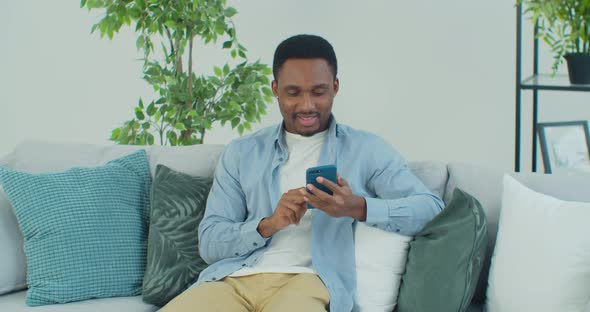 Afroamerican Man is Using Smartphone Sitting on Couch in Apartment Male Internet User Viewing Social