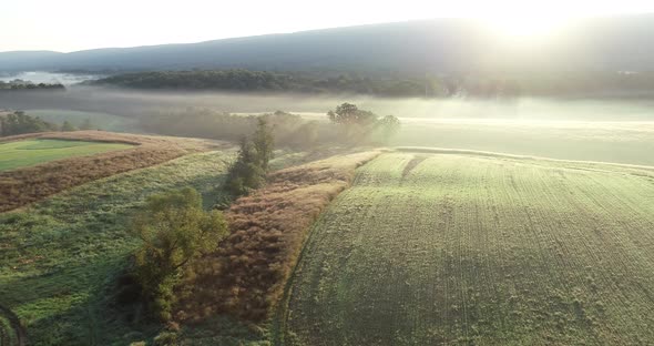 Aerial camera moving to the right showing farm fields, trees, mountains, and fog in the morning ligh