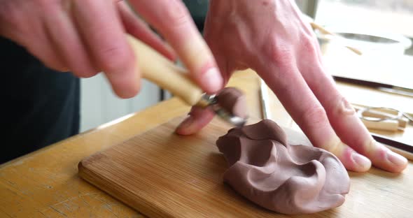 Close up on the hands of an artist carving and cutting brown modeling clay with a metal tool before