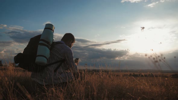 A Young Tourist Man with a Backpack Uses a Mobile Phone. In the Rays of the Setting Sun. Travel