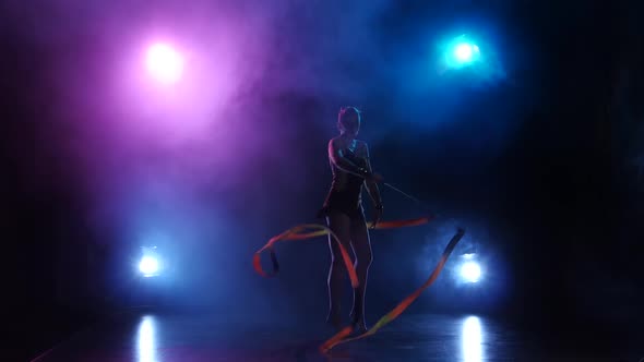 Gymnast with the Ribbon in His Hands Doing Acrobatic Moves. Black Background. Light Rear