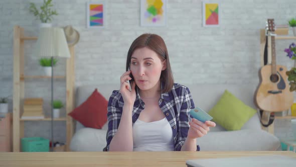 Irritated Young Woman with Credit Card in Hand Talking on the Phone in a Modern Apartment