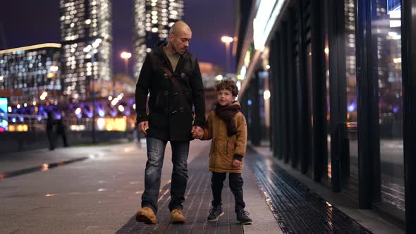 Father and Son are Walking Along a Blurry Evening Street Against the Background of City Lights