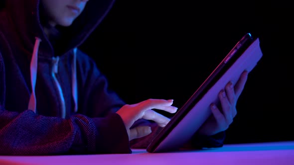 Young Woman in a Hood with a Tablet. Hacker Makes a Hack Through the Tablet. Blue and Red Light