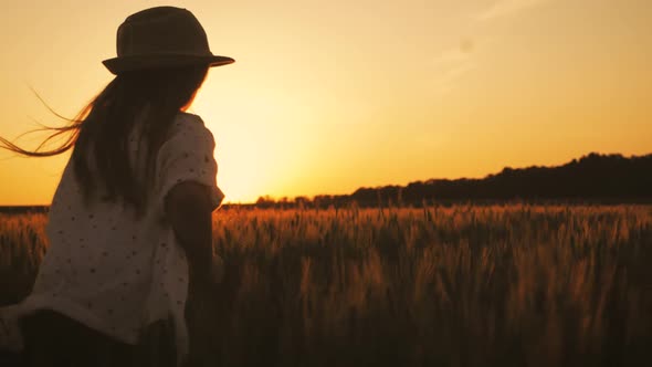 Pretty Child in the Hat Is Running Across the Wheat Field at Sunset