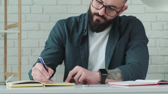 Close Up of Guy That Learn Online Courses Using a Tablet and Write Notice in His Notebook