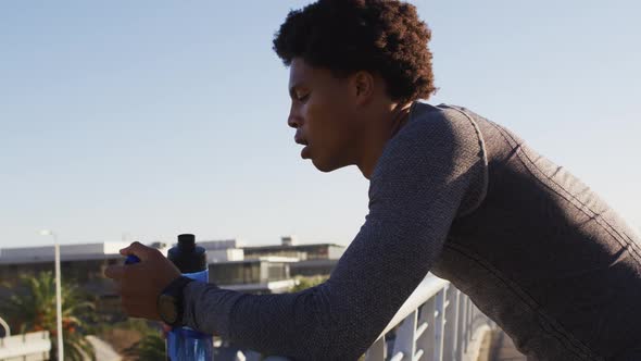 Fit african american man exercising in city, resting on footbridge, drinking from water bottle