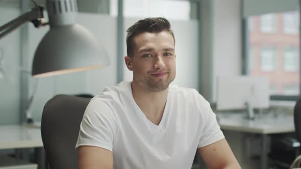 A Young Handsome Man in a White Tshirt Sits at an Office Desk and Winks Slow Motion