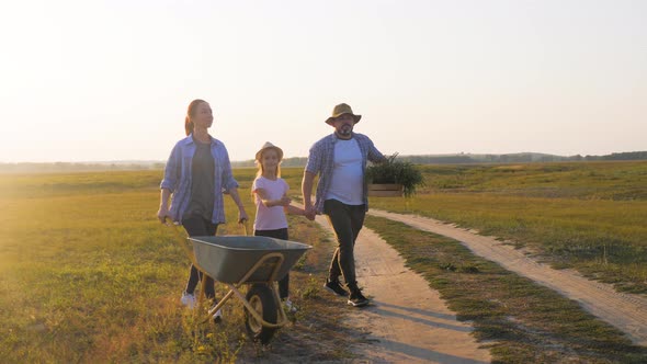 Young Family Having Fun Outdoors in Their Farm. Gardener Woman Pushing Wheelbarrow with Vegetables