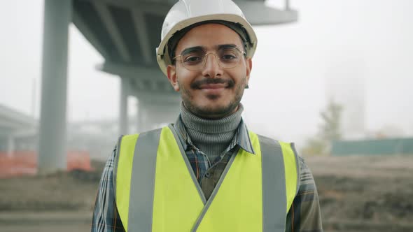 Portrait of Arab Man Wearing Helmet and Vest Smiling Standing Against Urban Construction Background