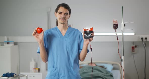 Male Nurse Holding Bag with Donor Blood and Red Rubber Heart Smiling at Camera
