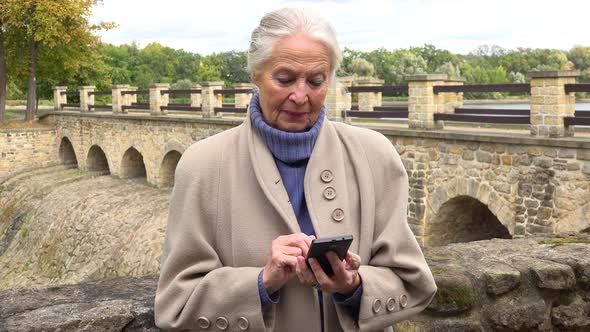 An Elderly Woman Leans Against the Stone Barrier of a Bridge and Works on a Smartphone