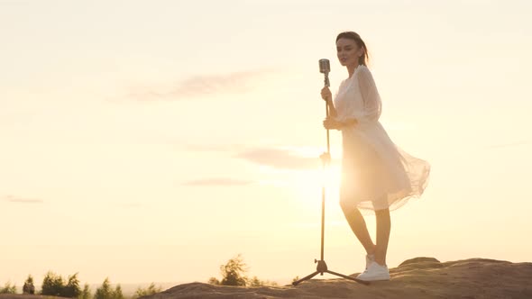 Beautiful Woman in White Dress on Top of Mountain on Edge of Abyss and Sings Song Into Shiny Retro