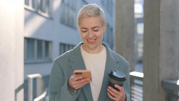 Happy Mature Lady Carrying Coffee and Using Mobile on Street