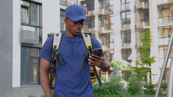 African Man Courier Delivery with Backpack Walks Street Watching in Smartphone