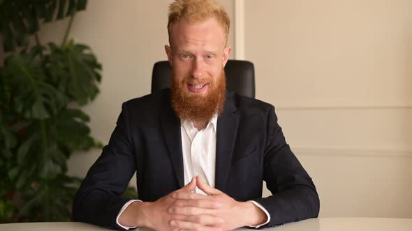 Redhaired Businessman in Suit Sitting at Desk Talks and Looks at the Camera