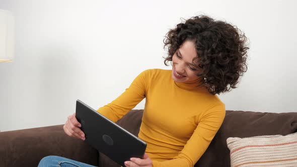 Curly Woman Emotionally Playing Video Game on Tablet Sitting on Couch at Home