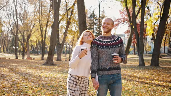 Young Positive Man and Woman Walking in Autumn Park Enjoying Warm Weather with Takeaway