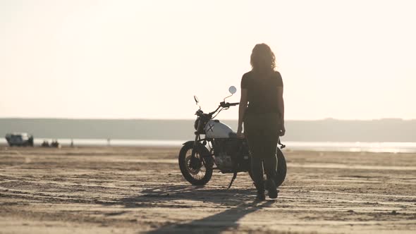 Beautiful Young Woman Riding an Old Cafe Racer Motorcycle on Desert at Sunset or Sunrise