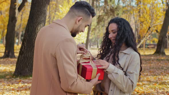 Girl Gives Gift to Beloved Boyfriend Young Guy Opens Red Box Unleashing Gold Ribbon Smiling Hispanic