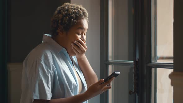 Excited Beautiful Afro Woman Using Smartphone While Standing Near Window Indoor