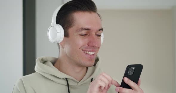 Closeup Portrait of Joyful Handsome Young Man Listening to Music in White Headphones and Surfing