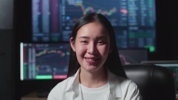 Asian Female Stock Market Broker Working On Computer And Smiling To Camera With Analysing Graphs