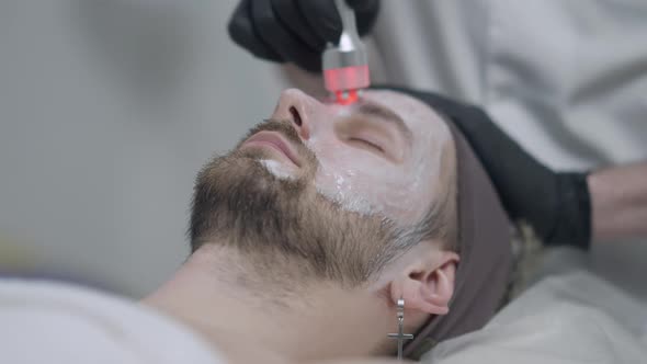 Closeup Face of Male Client Lying on Beauty Bed As Beautician in Gloves Doing Red LED Light