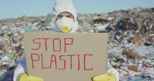 Man in Protective Suit Show Protest Sign "Stop Plastic" at Camera at Landfill