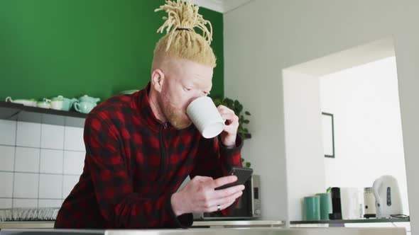 Thoughtful albino african american man with dreadlocks using smartphone and drinking coffee
