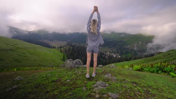 Young girl raised her hands up against background of clouds in mountains.