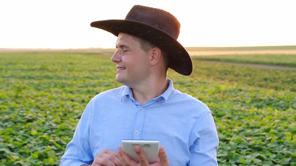 Farmer in a Hat Working on a Bean Field with a Tablet in His Hands on a Background of Sunlight