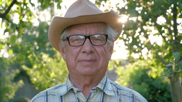 Old Caucasian Man With Glasses And Hat Portrait Against The Sunset In The Garden