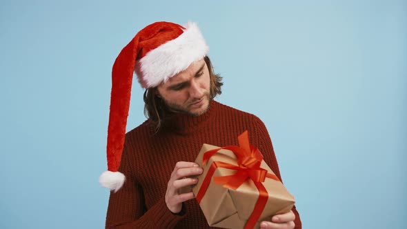 Man in Santa Hat is Holding Gift Box Shaking It Trying to Guess What is There Inside Smiling Posing