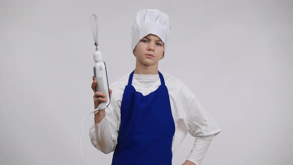 Portrait of Serious Caucasian Boy in Cook Uniform Turning on Electric Hand Blender Looking at Camera