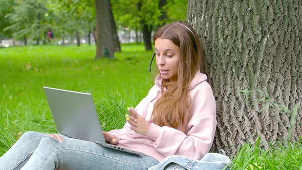 Smiling Woman with Laptop and Headset Having Online Conversation While Sitting in City Park.