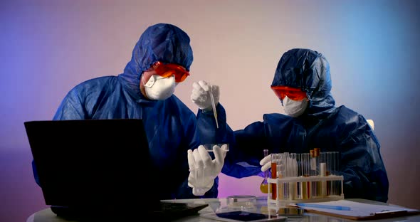 Two Virologist Doctors in a Protective Mask and Clothing Sit at a Table with a Laptop and Test Tubes