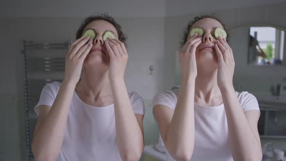 Two Cheerful Caucasian Young Slim Women Putting Cucumber on Eyes Laughing Taking Care of Skin Beauty