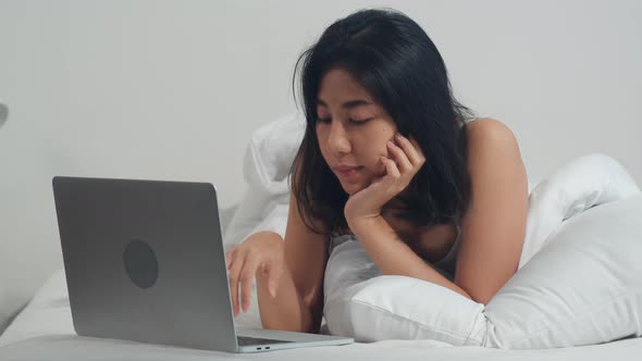 Young Asian woman using laptop checking social media feeling happy smiling while lying on bed