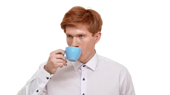 Young Caucasian Redhead Man Drinking Coffee From Blue Cup and Showing Ok on White Background in