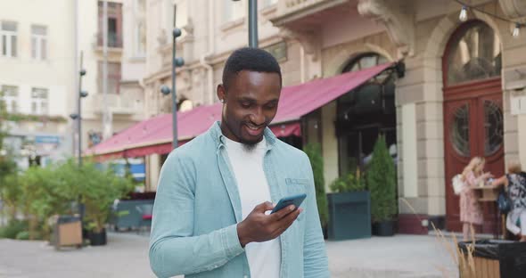 African American in Stylish Clothes Walking Near Cozy Cafe in Old City Part and Uses His Mobile