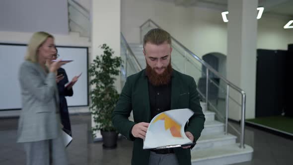 Concentrated Bearded Business Analyst Walking Analyzing Paperwork with Colleagues Passing in Office