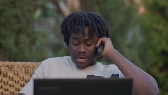 Concentrated Busy Young African American Man Talking on Smartphone Looking at Laptop Screen Sitting