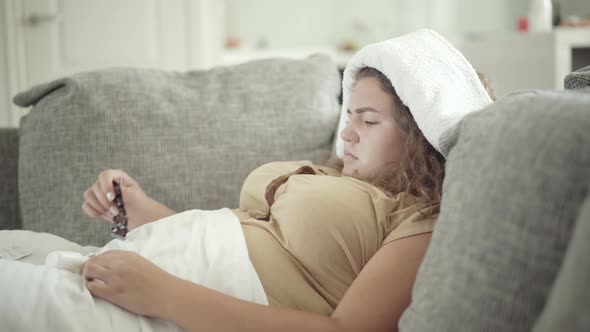 Concentrated Young Plump Woman Choosing Pills Lying on Couch with Towel on Head. Side View Portrait