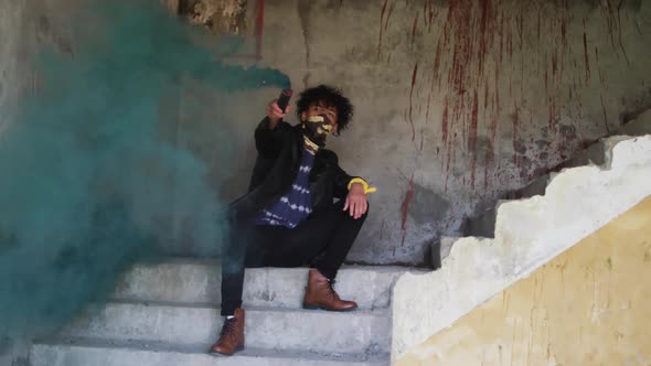 Mixed race man holding smoke grenade with green smoke sitting on steps in grungy warehouse