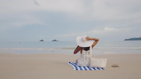 Woman In Sunhat Sitting On Beach Towel Looking Out To Sea